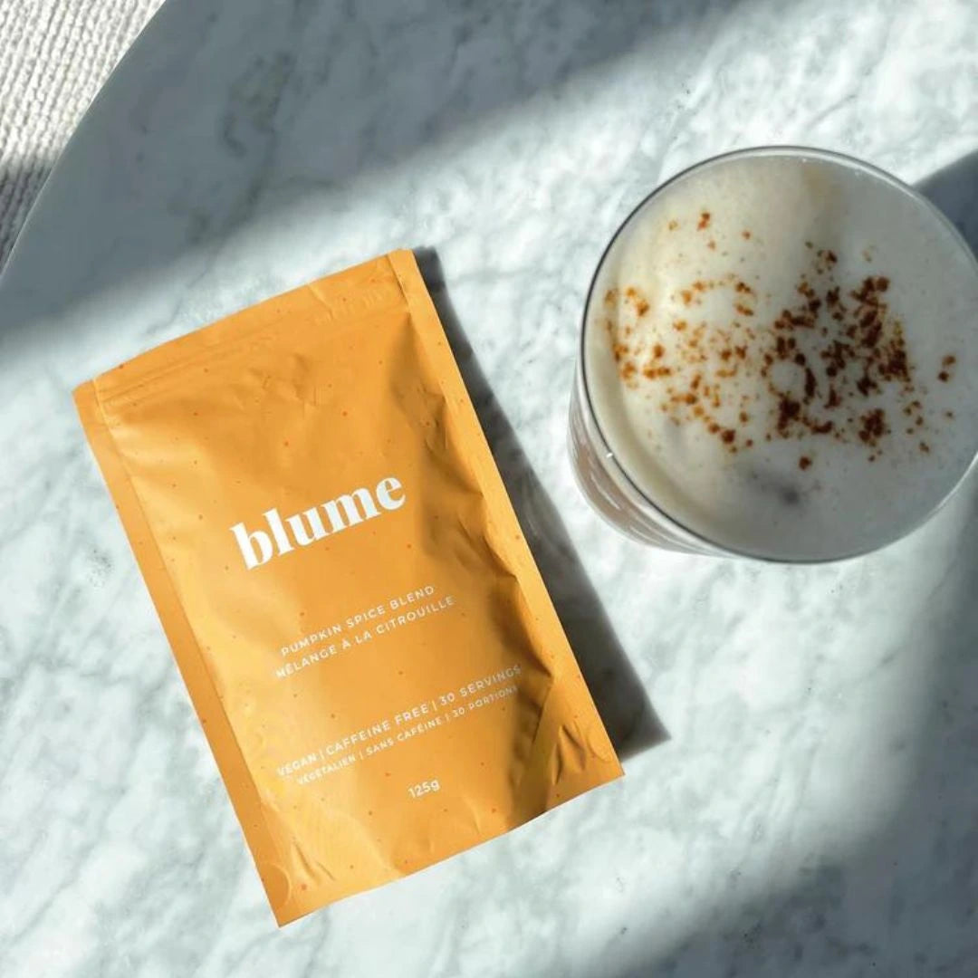 A packet of Blume Pumpkin Spice Blend lies on a marble surface next to a mug of frothy latte topped with a sprinkle of spice. Sunlight casts shadows across the scene.